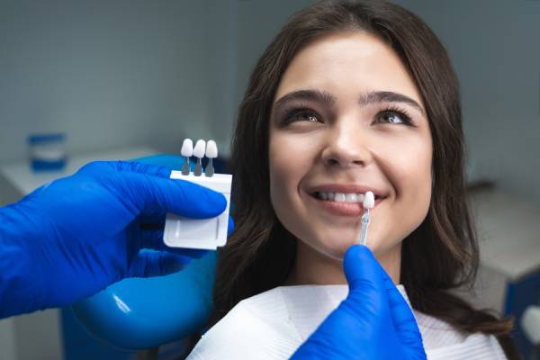 A woman receiving dental veneers at a dentists office, with gloved hands holding several differently shaded veneers in front of her